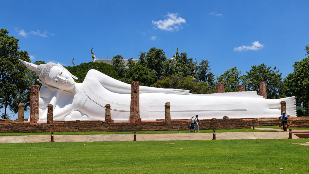 Stor hvid liggende Buddha under åben himmel med gamle murstenssøjler og besøgende ved Wat Khun Inthapramun.