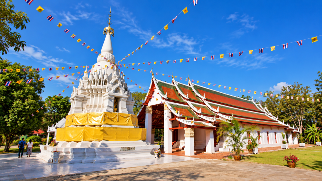 Hvid chedi med gult klæde ved siden af en tempelbygning med rødt og grønt tag ved Wat Phra Borommathat Worawihan.